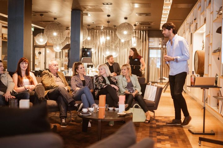 A group of people seated on couches in a modern lounge, listening to a man standing and speaking.