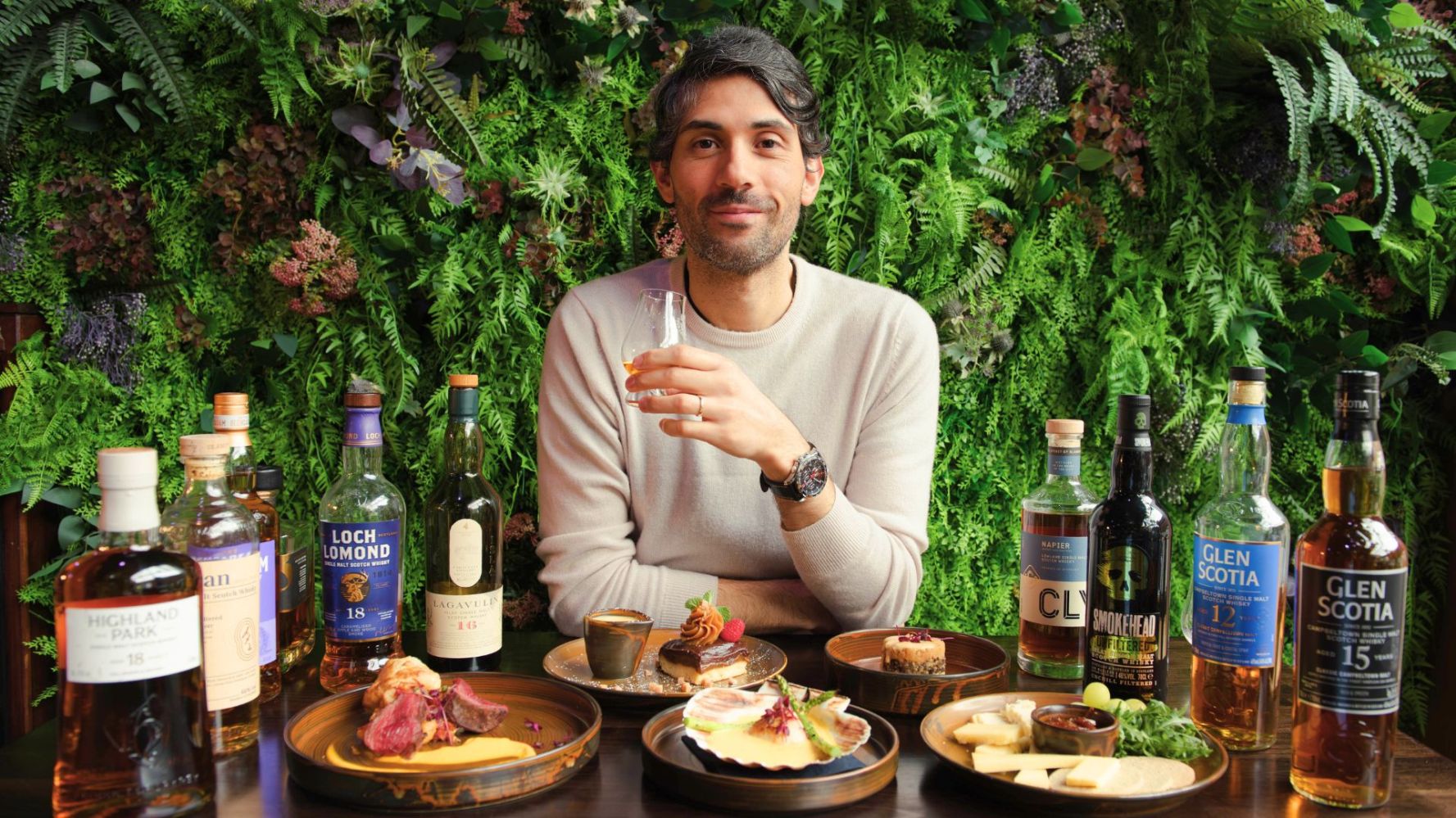 Camilo Gomez Founder of Once Upon a Whisky sitting with whisky bottles and assorted Scottish dishes in a lush green setting.