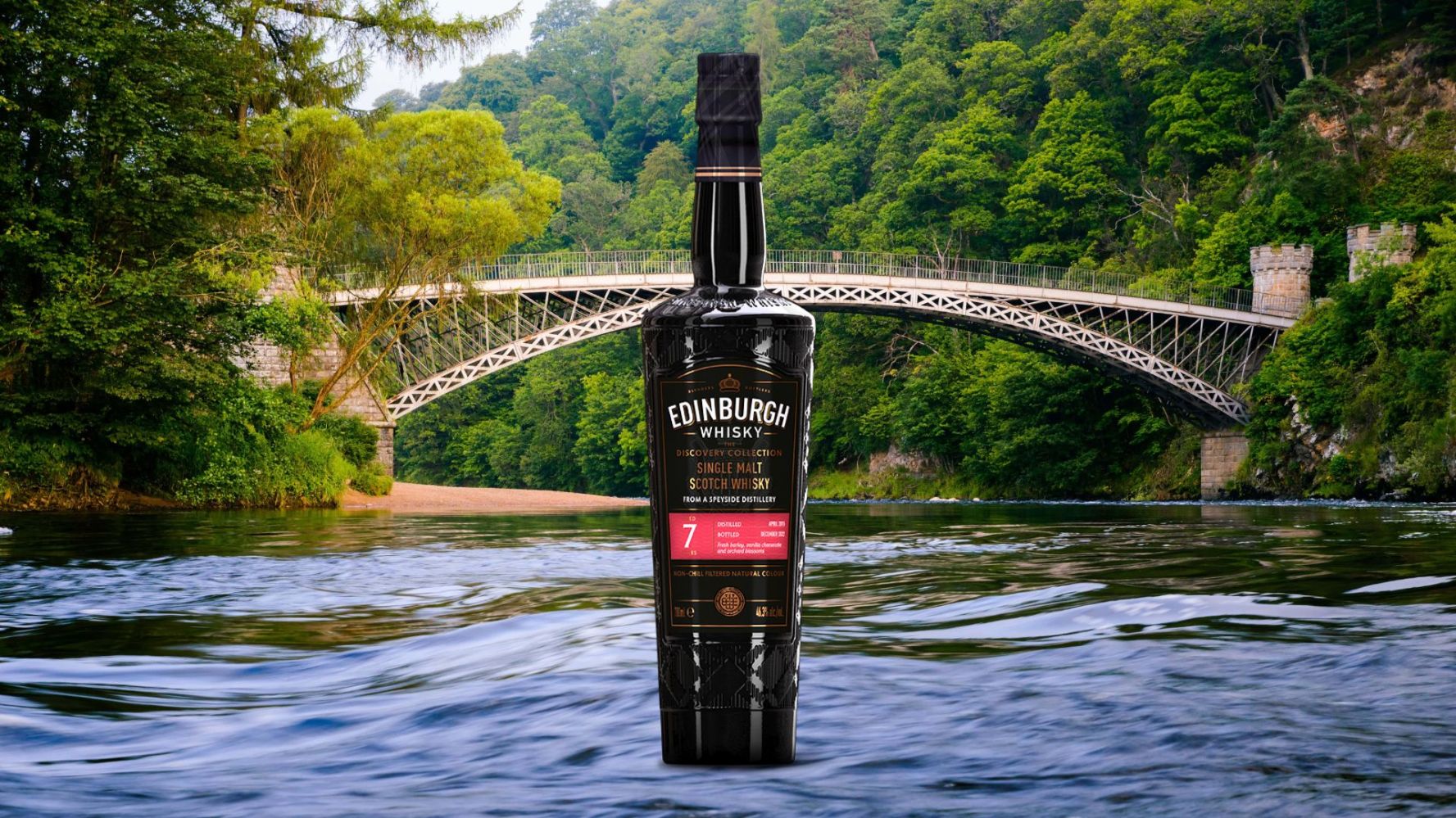 Whisky bottle in front of a scenic bridge over a river with lush greenery.