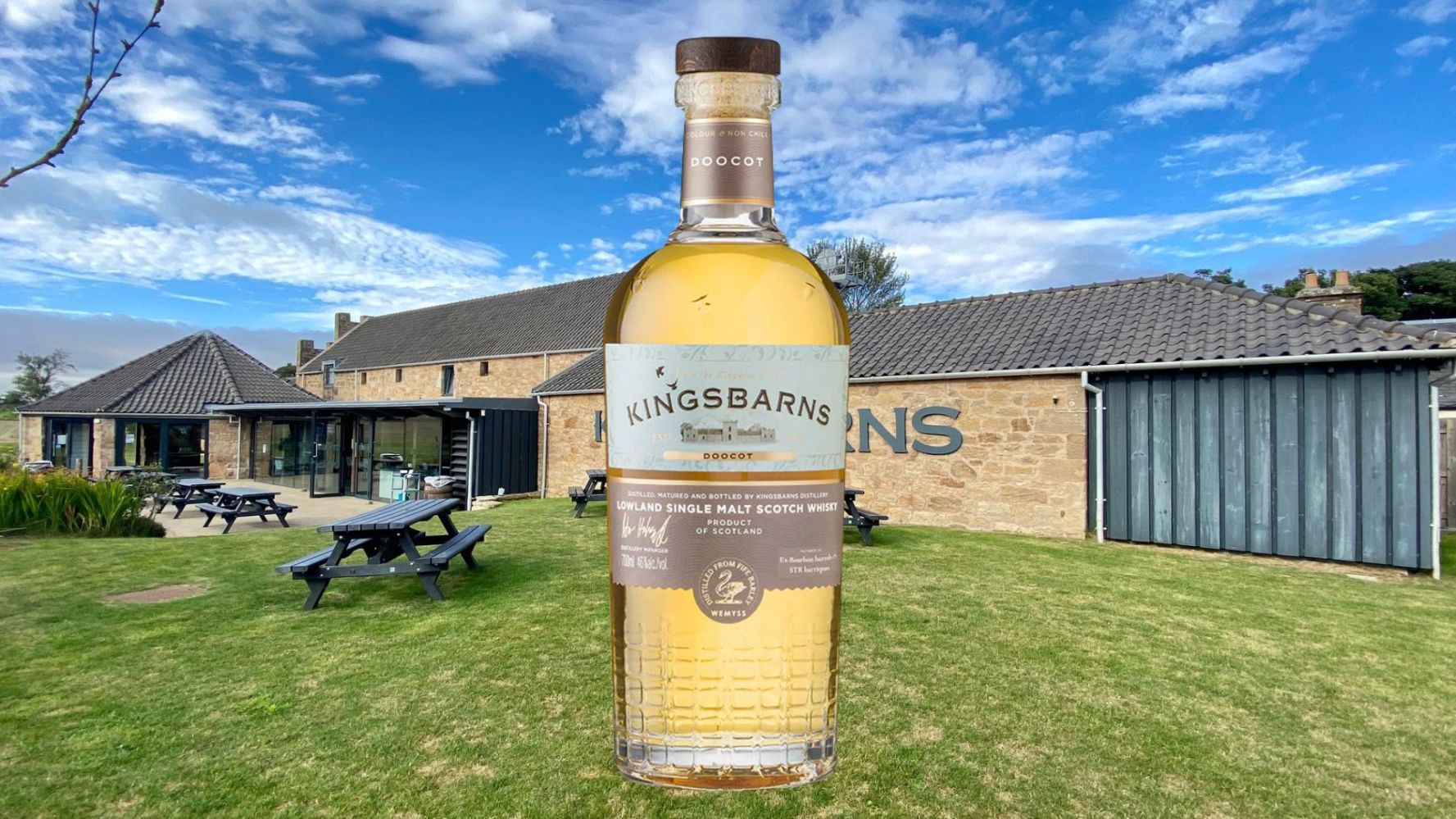 Kingsbarns whisky bottle overlaid on a distillery with picnic tables and blue sky.