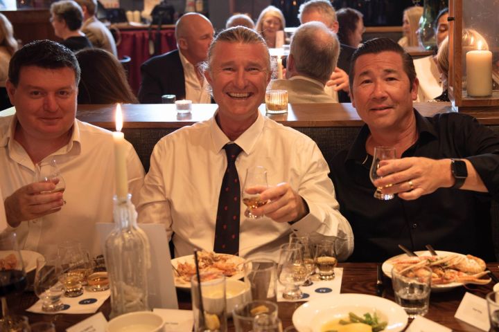 Three men in formal wear at a restaurant table, smiling and holding drinks.