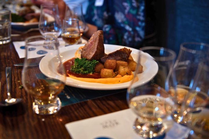 Plate of meat, potatoes, and broccoli on a table with glasses of whiskey.