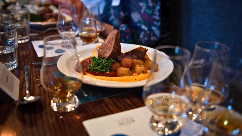 Plate of meat, potatoes, and broccoli on a table with glasses of whiskey.