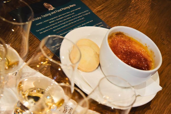 Plate with crème brûlée and biscuits, glasses of whisky, and menu on wooden table.