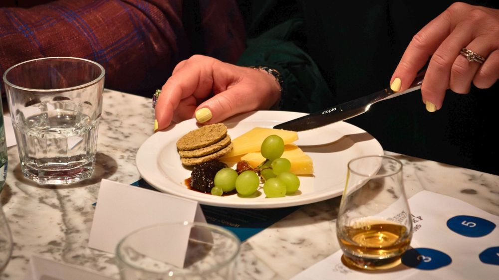 Hand slicing cheese with crackers, grapes, and figs on a plate next to glasses of water and whiskey.
