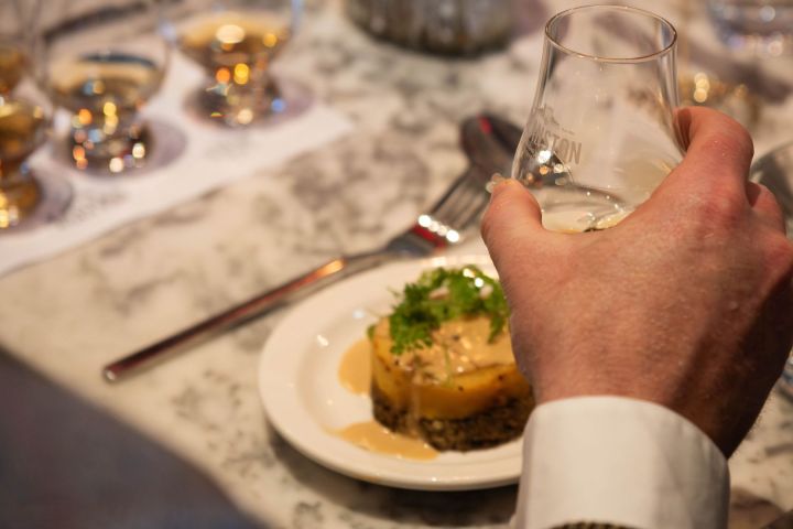 Person holding a glass of whisky over a gourmet dish on a table with whisky glasses.