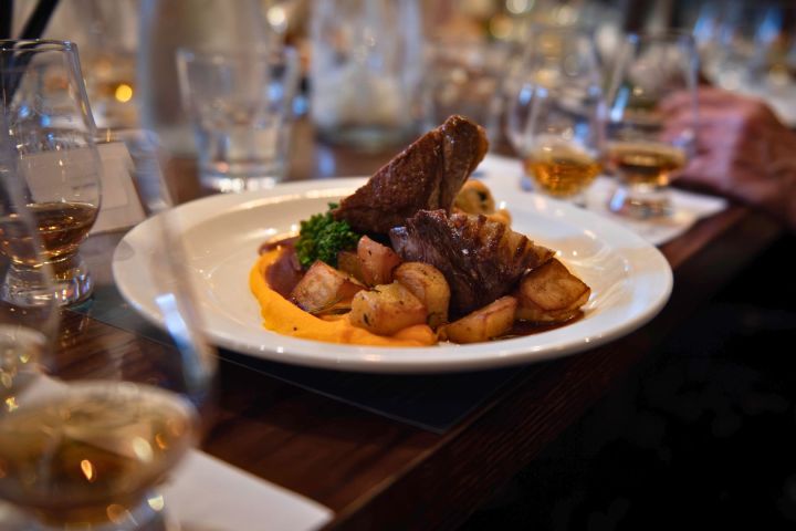 Plate of beef with roasted potatoes and puree on a table set with glasses of whiskey.