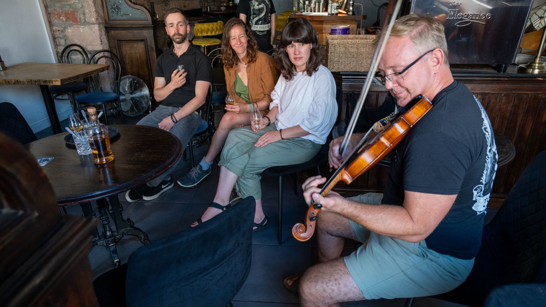 Man plays violin in a cozy room with three people sitting and watching nearby.