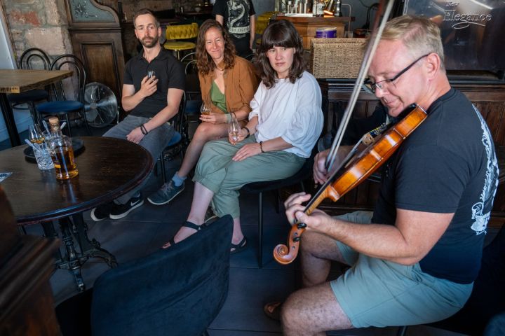 Man plays violin in a cozy room with three people sitting and watching nearby.