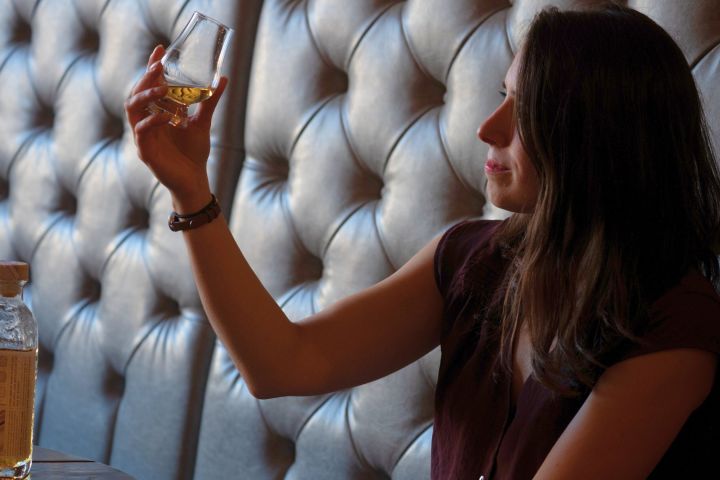 Woman seated, holding and examining a glass of whiskey with bottle nearby.