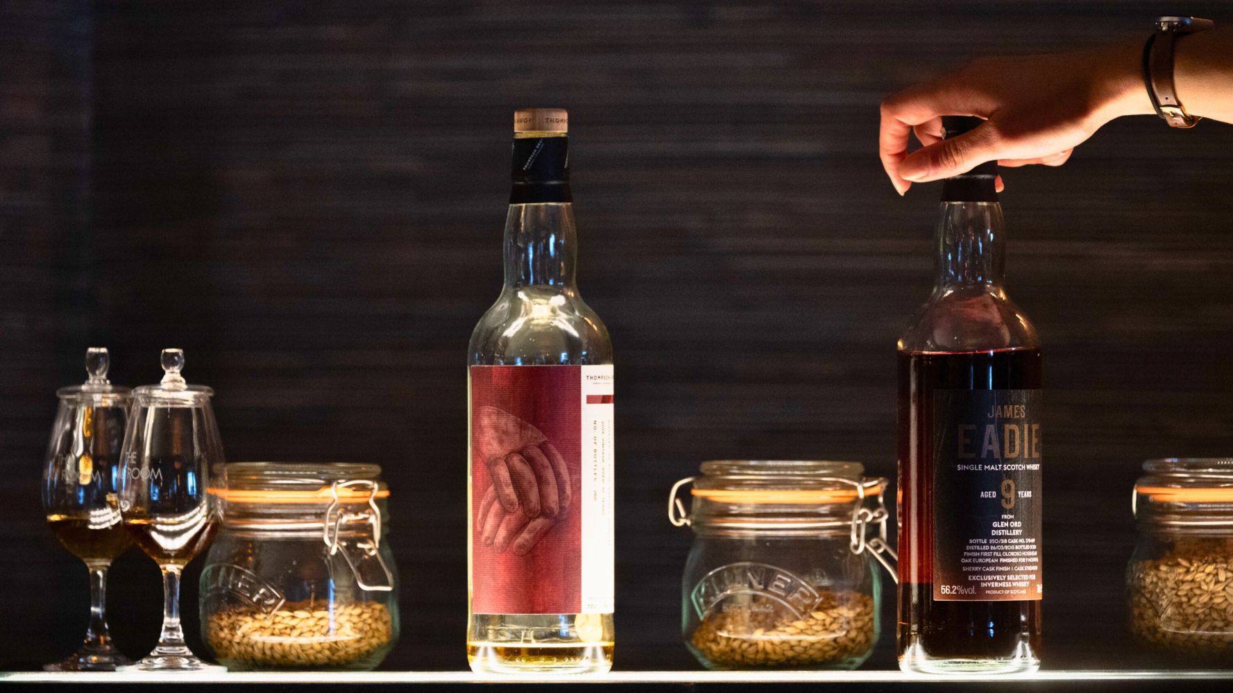 Hand reaches for a whiskey bottle among jars and glasses on a bar shelf.