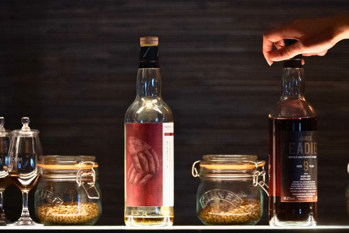 Hand reaches for a whiskey bottle among jars and glasses on a bar shelf.