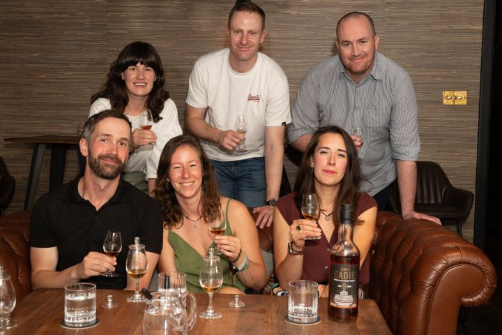 Six people smiling, seated on couches, holding glasses, with a bottle on the table.