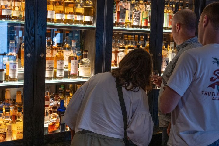Three people observe a display of assorted whiskey bottles in glass cabinets.