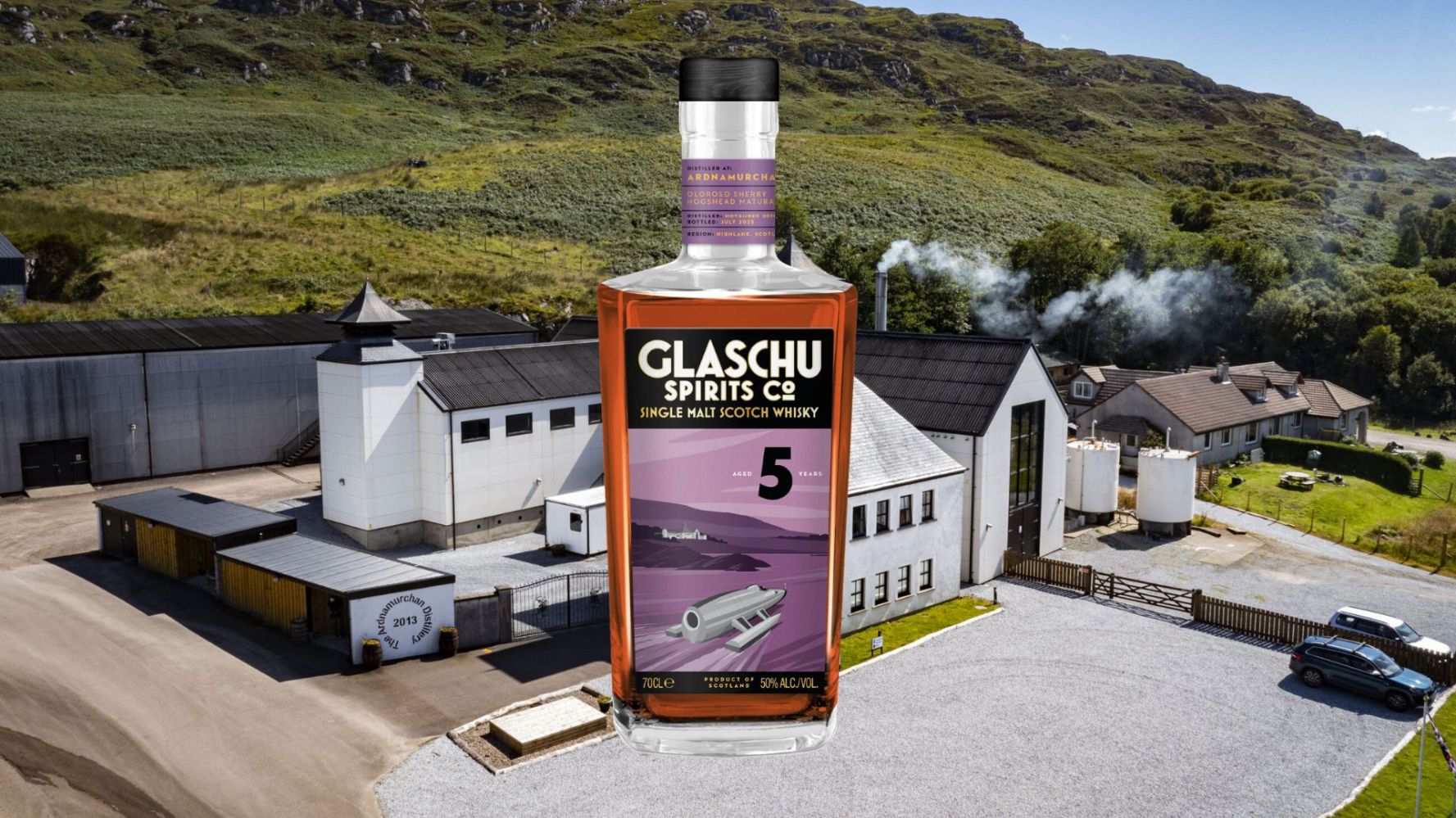 Whisky bottle in front of distillery building in scenic countryside.