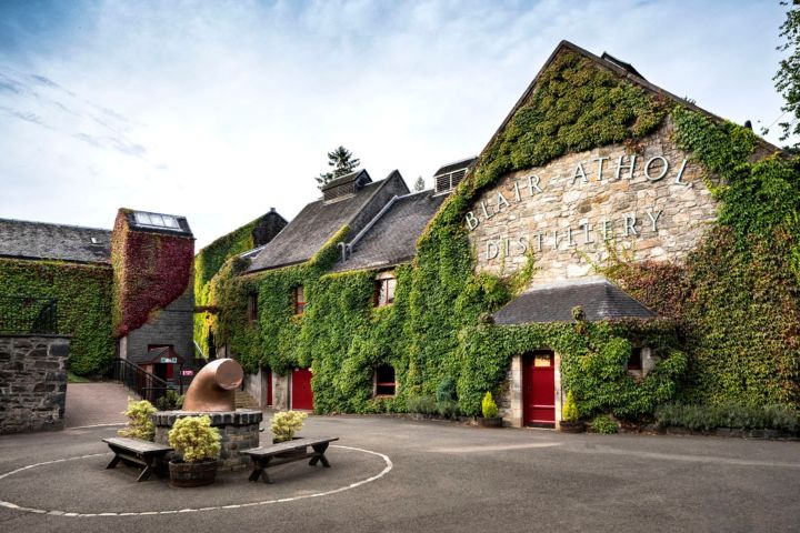 Ivy-covered distillery building with red doors, benches, and a copper pot sculpture in front.