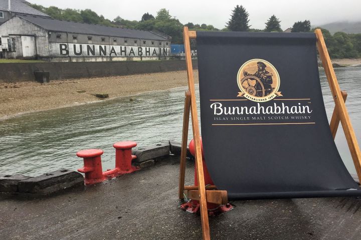 Large deck chair with Bunnahabhain logo by the waterfront, distillery in the background.