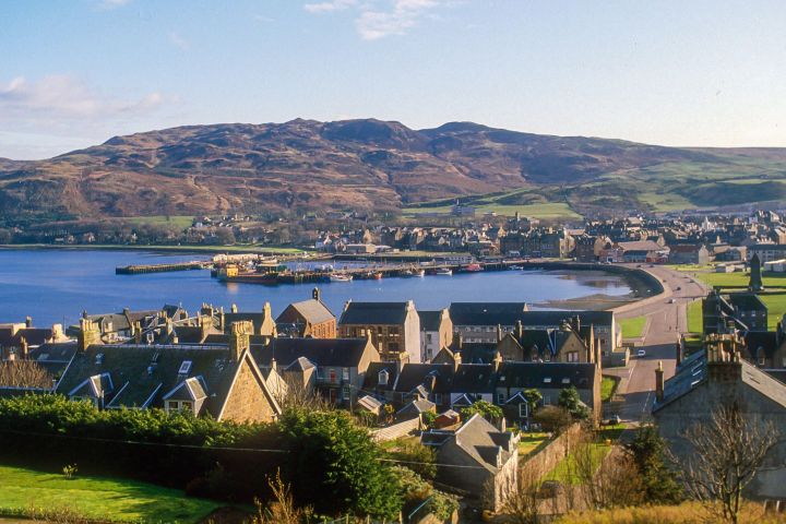 Coastal town with rooftops in foreground and hills in the background on a sunny day.