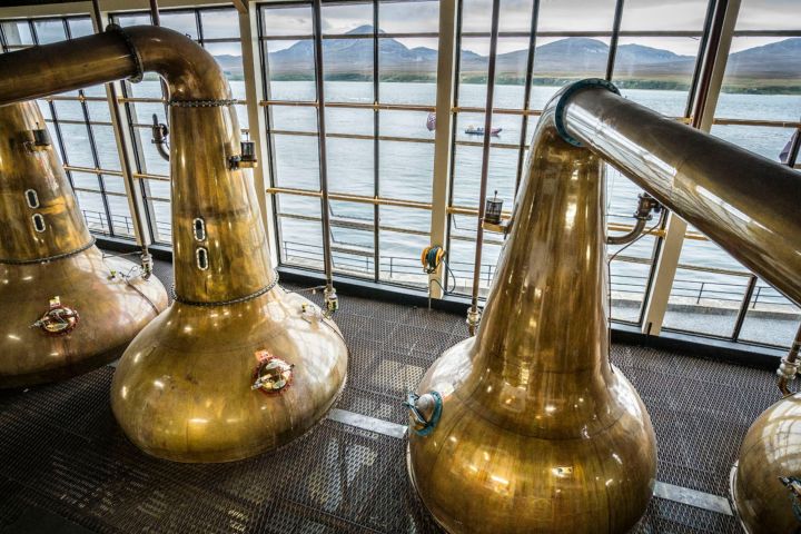 Copper stills in a distillery with large windows overlooking a scenic lake and mountains.