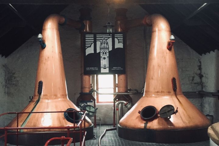 Copper distillation tanks inside a distillery with dark ceiling and light streaming through a window.