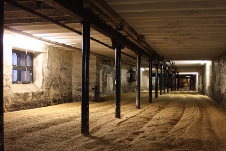 Industrial room with stone walls and wooden floor covered in grain, supported by black metal columns.