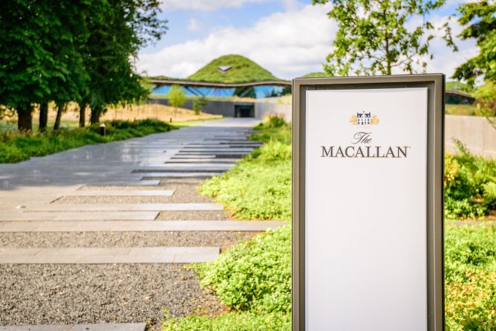 Entrance path to The Macallan distillery with greenery and modern building in background.