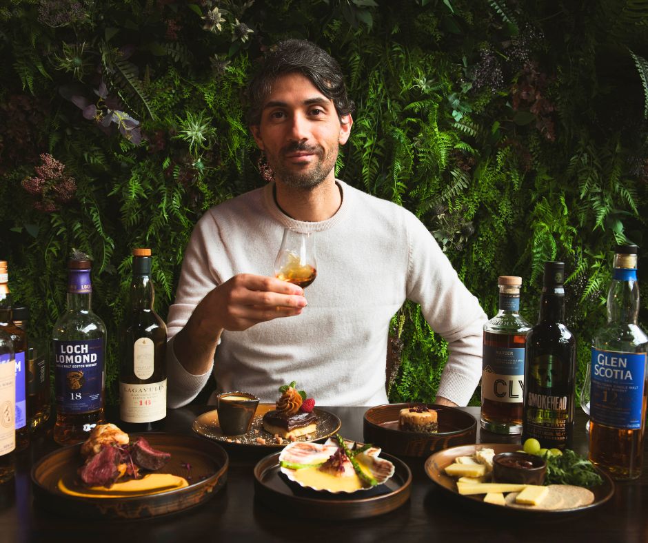 A man toasting with a whisky in his hand on a table full of food and whisky