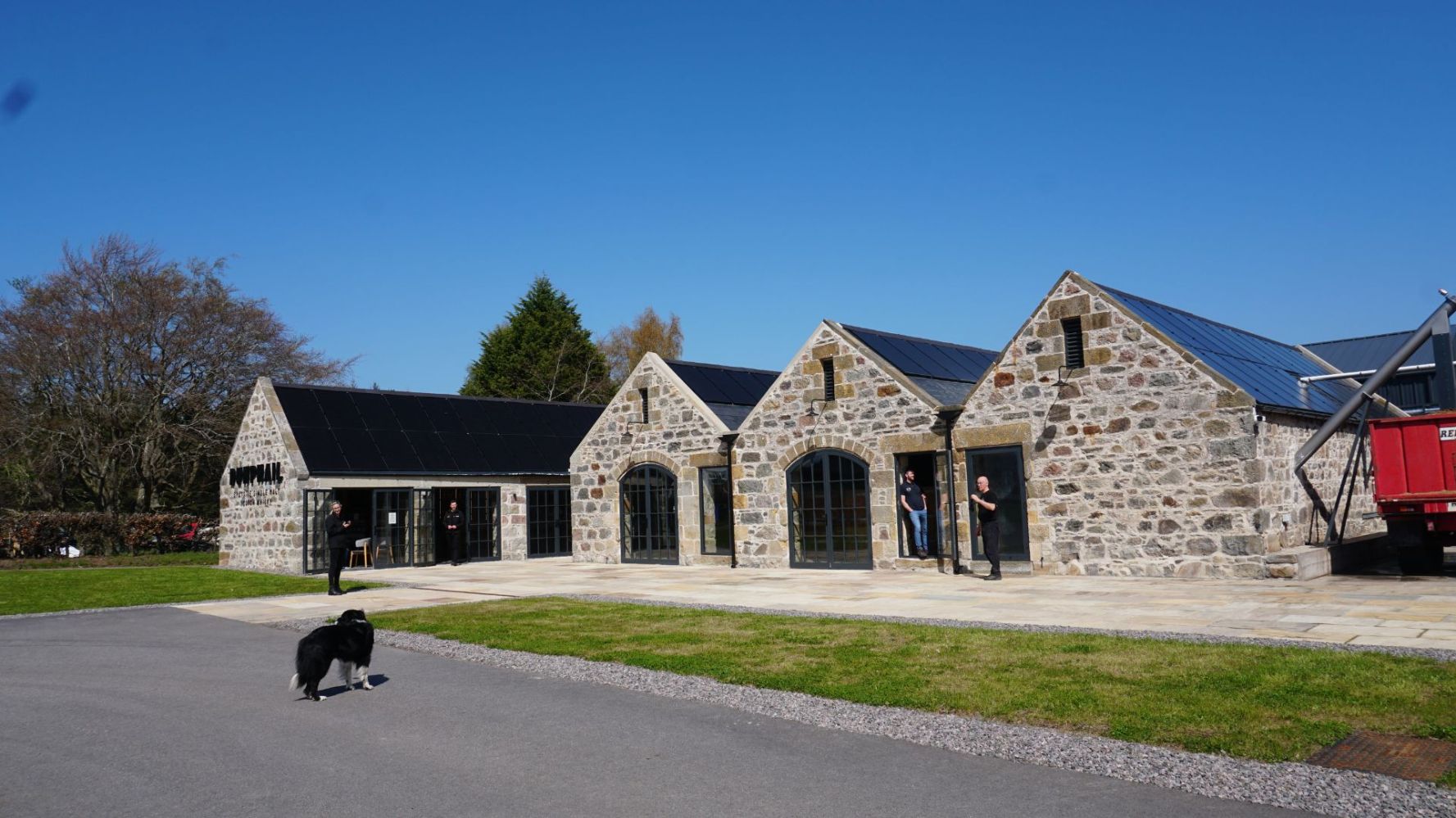 Stone building with arched windows, people, and a dog on a paved path under a clear blue sky.