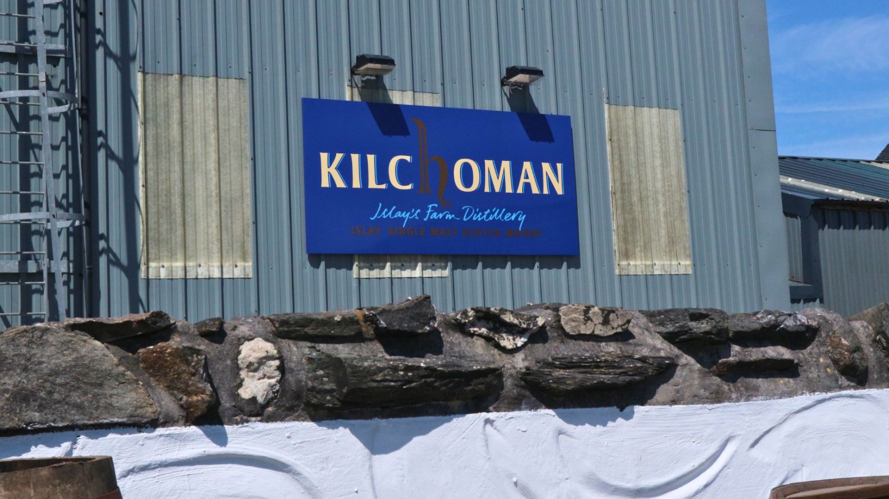 Kilchoman sign on corrugated metal building with stone wall in foreground.
