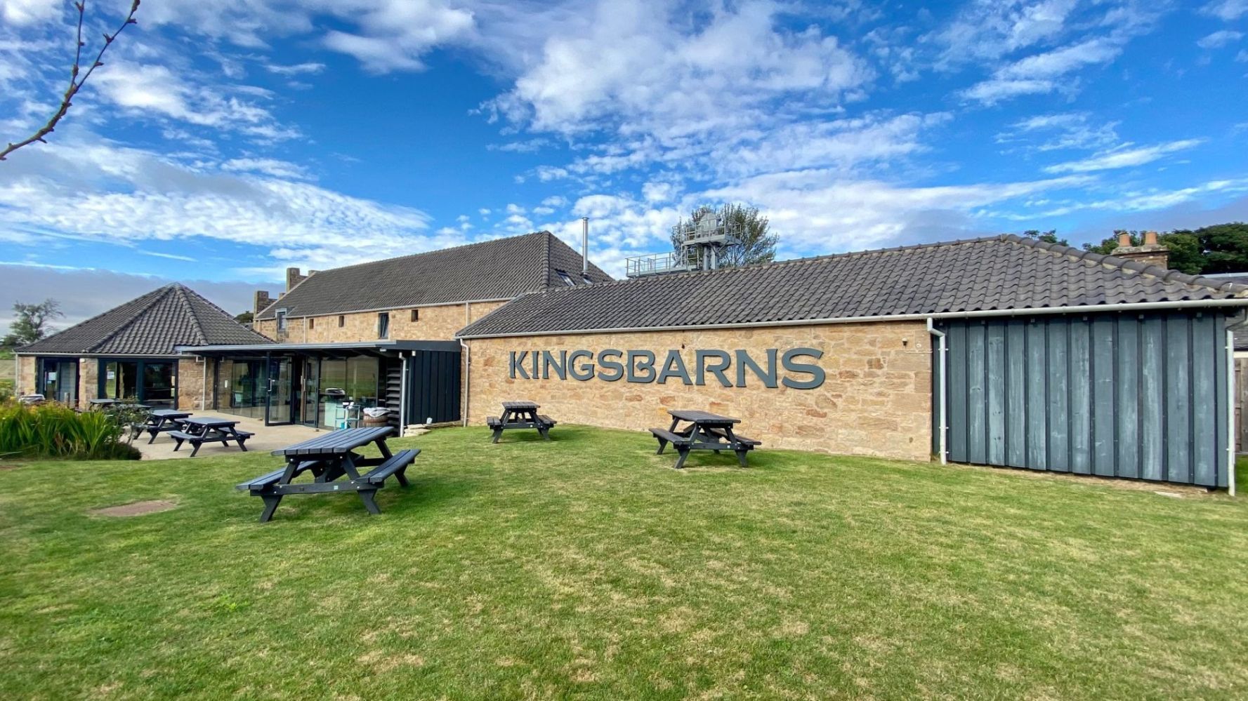Exterior of building with 'KINGSBARNS' sign and picnic tables on grass.