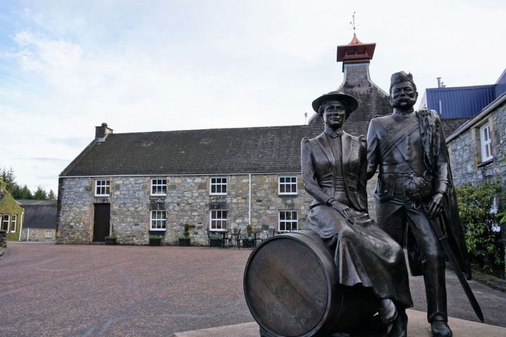Statues of a man and woman on a barrel with the Glenfiddich Distillery behind.