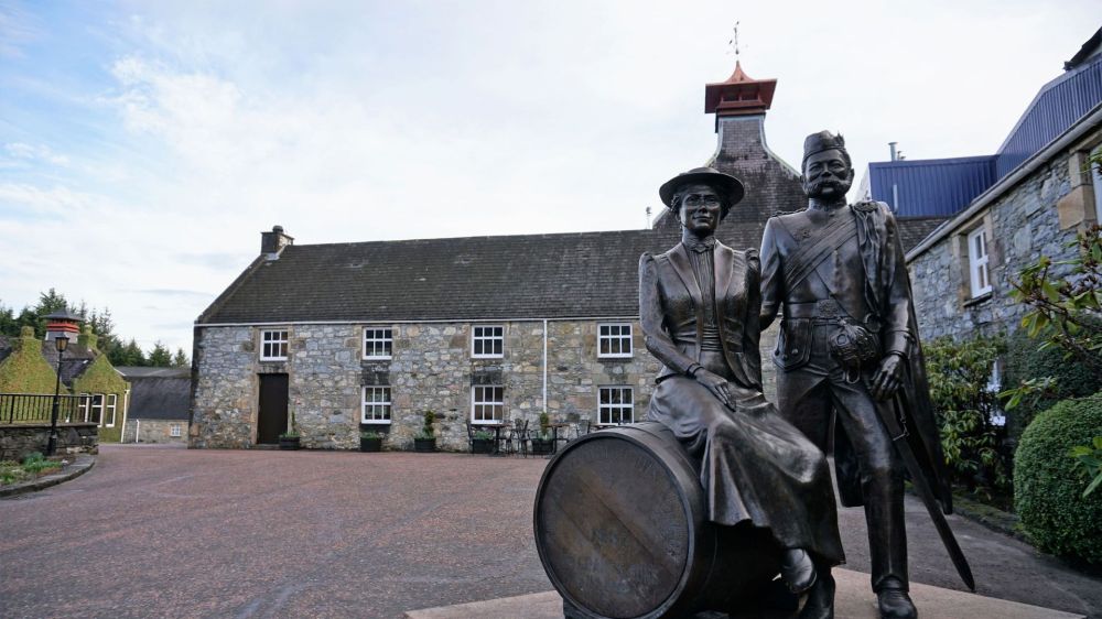 Statues of a man and woman on a barrel with the Glenfiddich Distillery behind.