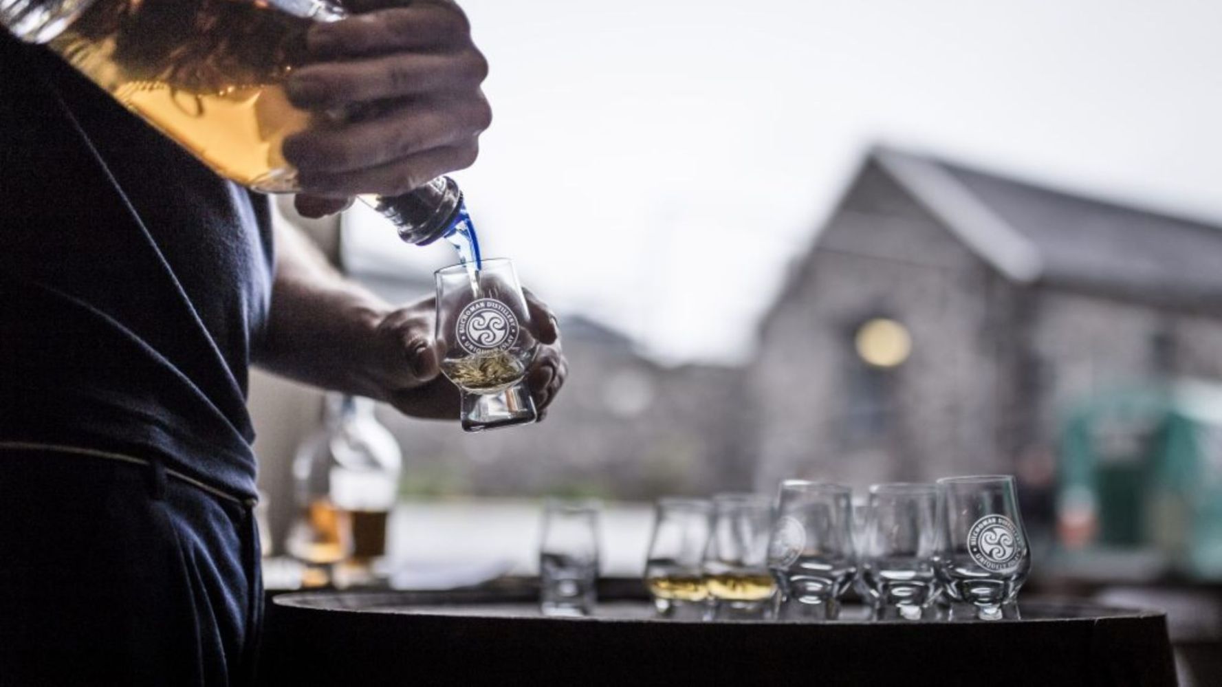 Person pouring whiskey into branded glass outdoors.